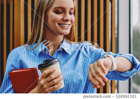 Stylish hipster girl holding book and coffee to go in hand while managing clock on wristwatch 108992381