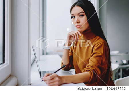 attractive confident businesswoman looking at camera making notes and plannings near window attractive confident businesswoman looking at camera making notes and plannings near window 108992512