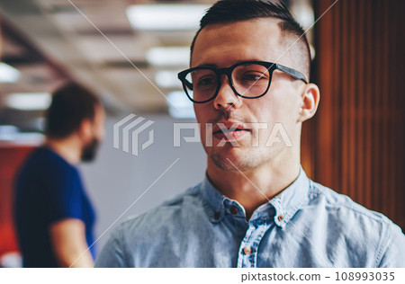 Serious young man in eyeglasses standing in office and communicating with colleague Serious young man in eyeglasses standing in office and communicating with colleague 108993035