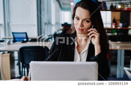 serious female employee sitting at desktop with notebook making cellphone call for banking 108993134