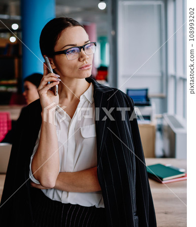 Serious female in eyewear looking away in office making mobile phone call for job Serious female in eyewear looking away in office making mobile phone call for job 108993202