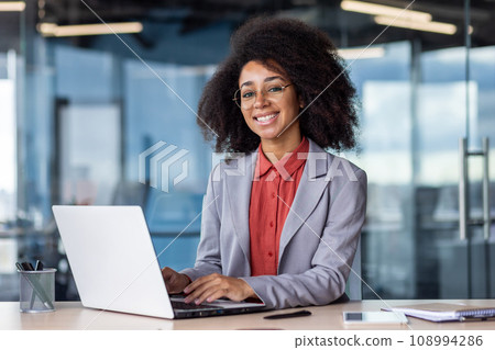 Portrait of young beautiful smiling businesswoman at workplace inside office, woman in business suit smiling and looking at camera, female financial worker using laptop at work. Portrait of young beautiful smiling businesswoman at workplace inside office, woman in business suit smiling and looking at camera, female financial worker using laptop at work. 108994286