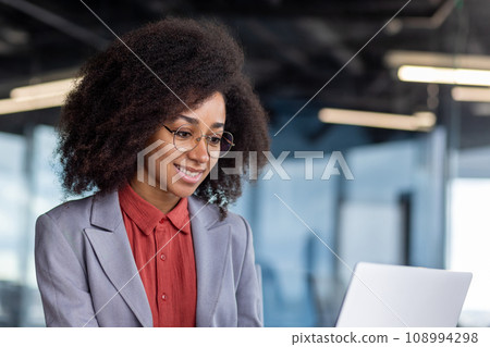 Young beautiful successful business woman close up smiling looking at laptop screen, female worker in business suit with curly hair is reading contentedly, woman is happy results of achievement. Young beautiful successful business woman close up smiling looking at laptop screen, female worker in business suit with curly hair is reading contentedly, woman is happy results of achievement. 108994298
