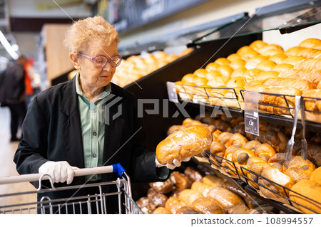 elderly woman with glasses is shopping in the bread department of a supermarket 108994557