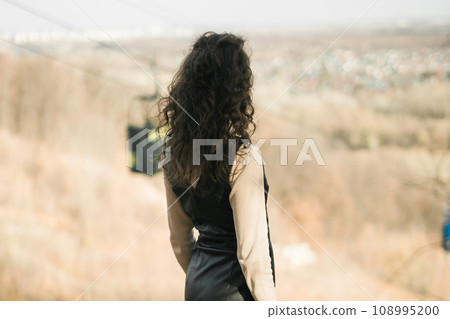 Back view Girl in fall park against background of cable car. Woman with long curly hair. Beautiful sunlight in the forest. Hair care. Millennial Generation and youth. 108995200