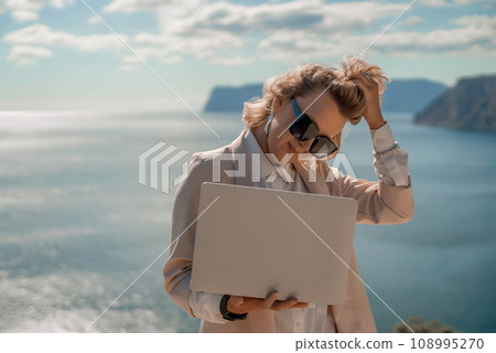 Freelance women sea. She is working on the computer. Good looking middle aged woman typing on a laptop keyboard outdoors with a beautiful sea view. The concept of remote work. Freelance women sea. She is working on the computer. Good looking middle aged woman typing on a laptop keyboard outdoors with a beautiful sea view. The concept of remote work. 108995270