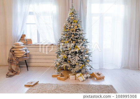Snow-covered Christmas tree with gifts and toys in the interior of a room with windows Snow-covered Christmas tree with gifts and toys in the interior of a room with windows 108995308