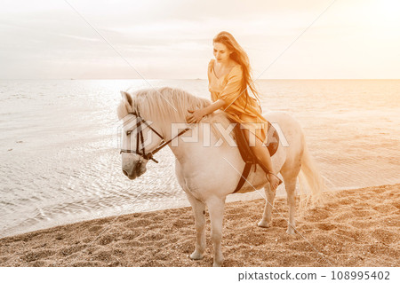 A woman in a dress stands next to a white horse on a beach, with the blue sky and sea in the background. A woman in a dress stands next to a white horse on a beach, with the blue sky and sea in the background. 108995402
