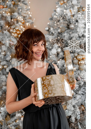 Woman Christmas tree gift. Happy romantic brunette girl in black dress standing on the background of the Christmas tree. Cheerful lady was surprised by the gift after opening the gift box. Merry Woman Christmas tree gift. Happy romantic brunette girl in black dress standing on the background of the Christmas tree. Cheerful lady was surprised by the gift after opening the gift box. Merry 108995464