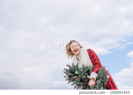 Blond woman holding Christmas tree by the sea. Christmas portrait of a happy woman walking along the beach and holding a Christmas tree in her hands. Dressed in a red coat, white suit. 108995465