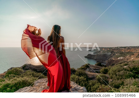 Woman sunset sea red dress, back view a happy beautiful sensual woman in a red long dress posing on a rock high above the sea on sunset. 108995466