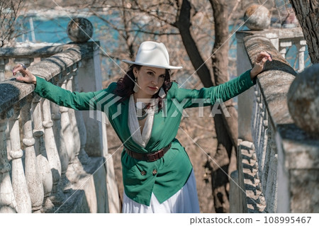 Woman walks around the city, lifestyle. Happy woman in a green jacket, white skirt and hat is sitting on a white fence with balusters overlooking the sea bay and the city. Woman walks around the city, lifestyle. Happy woman in a green jacket, white skirt and hat is sitting on a white fence with balusters overlooking the sea bay and the city. 108995467