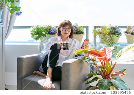 Middle aged happy woman sitting at home in chair with cup of smartphone 108995496
