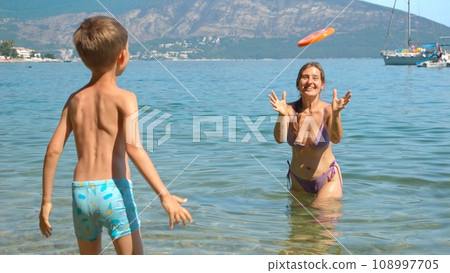 Cheerful boy with mother throwing and catching frisbee disk while relaxing in the calm sea water at beach. Family holiday, vacation and fun summertime of children and parents. Cheerful boy with mother throwing and catching frisbee disk while relaxing in the calm sea water at beach. Family holiday, vacation and fun summertime of children and parents. 108997705