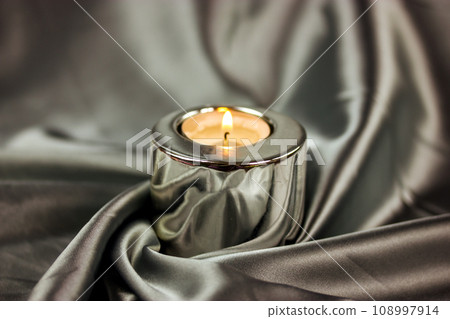 A burning candle in a metal candle holder on a table covered with a silver silk tablecloth. Luxurious stylish decor for modern interior A concept of tranquility, relaxation, meditation, self-knowledge A burning candle in a metal candle holder on a table covered with a silver silk tablecloth. Luxurious stylish decor for modern interior A concept of tranquility, relaxation, meditation, self-knowledge 108997914