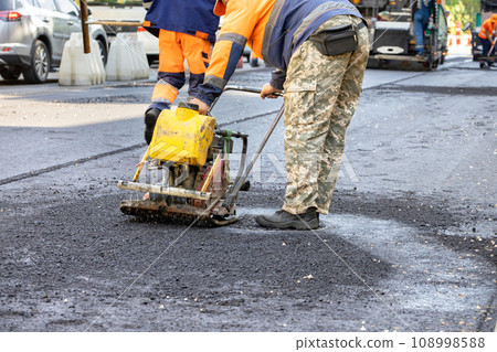 A road worker compacts fresh asphalt with a gasoline vibrating plate around a sewer manhole on the road. A road worker compacts fresh asphalt with a gasoline vibrating plate around a sewer manhole on the road. 108998588