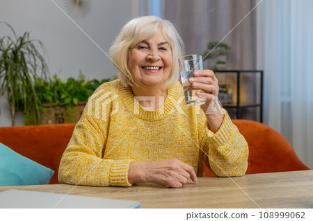 Thirsty senior old woman sit at home holding glass of natural aqua make sips drinking filtered water Thirsty senior old woman sit at home holding glass of natural aqua make sips drinking filtered water 108999062