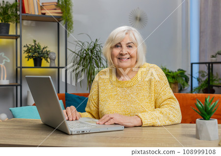 Happy senior old woman grandmother sitting on sofa at table working on laptop pc at office home room 108999108