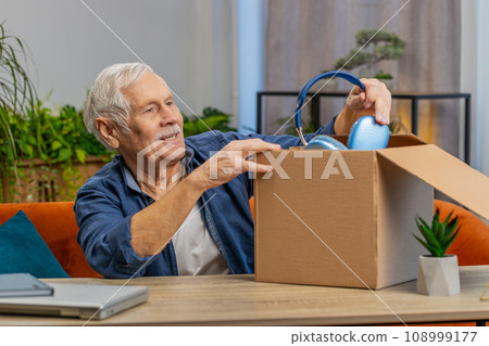 Happy senior man shopper unpacking headphones cardboard box delivery parcel online shopping purchase 108999177