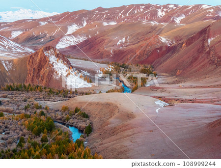 Scenic motley autumn landscape with yellow larch trees on frozen red hills, blue river and mountain range. Vivid autumn colors in mountains. Valley of the Kokorya River in Altai in the early morning. 108999244