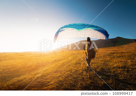 male paraglider takes off from a yellow field with a blue parachute against the backdrop of hills and small mountains. Paragliding 108999417