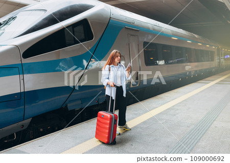 Young refugee woman waiting train on station platform, she lost and using smart phone. Traveler. 30s Women with suitcase walking at railroad station platform. Travel to vacation by train, full body Young refugee woman waiting train on station platform, she lost and using smart phone. Traveler. 30s Women with suitcase walking at railroad station platform. Travel to vacation by train, full body 109000692