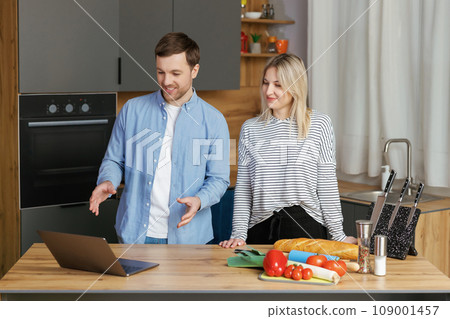 Pleasant family couple standing near big wooden table in modern kitchen, looking at laptop screen. Happy young couple starting to prepare lunch. 109001457