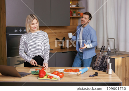 Beautiful young couple is talking and smiling while cooking healthy food in kitchen at home. Man is drinking cup of coffee and talking with woman. 109001730