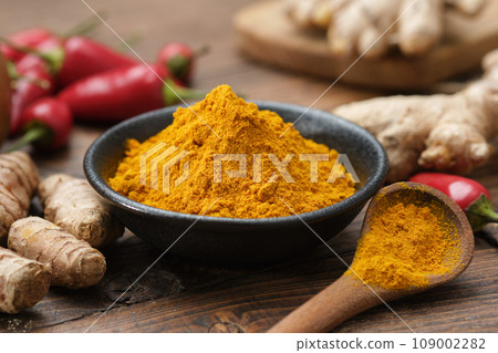 Bowl of curcuma ground to powder and whole turmeric roots. Red chilli pepper pods and ginger roots on background. Ingredients for cooking and Ayurveda. 109002282