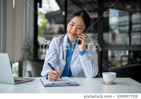 A doctor is talking on the phone with her patient while working in her hospital office. A doctor is talking on the phone with her patient while working in her hospital office. 109007789