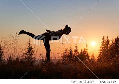 Young woman doing warrior 3 pose at fresh air. Silhouette of sporty female practicing yoga in mountains at sunrise. Exercising outdoor at early morning. 109013136