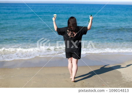A girl in a black dress and a bottle of champagne is standing on the beach 109014813
