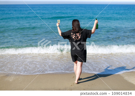 A girl in a black dress with champagne and a glass is standing on the seashore 109014834
