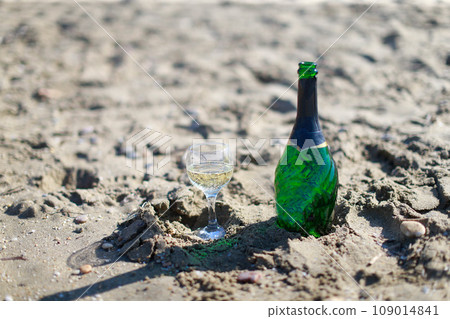 A bottle of champagne with a poured glass stands on the sand A bottle of champagne with a poured glass stands on the sand 109014841