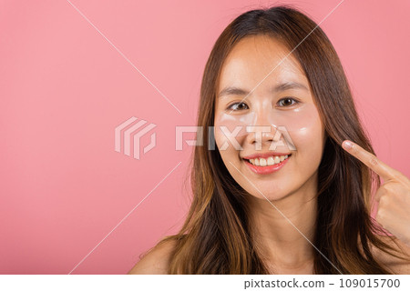 Eye treatment. Asian beautiful young woman smiling point finger to cosmetic patches gel under eyes, Female with natural makeup hydrogel eye patch on face, isolated on pink background, skincare 109015700
