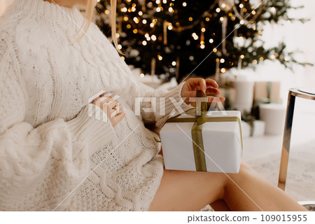 pregnant woman wearing knitted dress staying in the hall, on the background there is christmas tree with lights and toys on it Pregnant woman holding a little gift in her hands 109015955