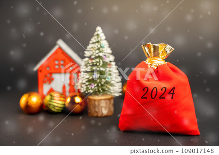 Red gift bag 2024, Christmas tree and miniature house. Merry Christmas and Happy New Year! Xmas concept. Festive winter holiday. Snowfall 109017148