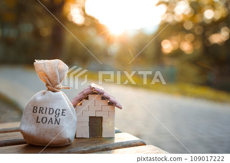 Bridge loan concept - is a short-term loan secured by real estate.Money bag and miniature house in the park in the sunset light. Real estate and finance 109017222