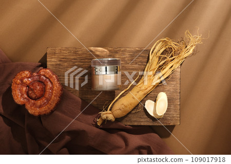 On a wooden podium, a cosmetic jar decorated with root and slices of ginseng, lingzhi mushroom and brown cloth on brown background. Concept for cosmetics extracted from rare herbs. Top view On a wooden podium, a cosmetic jar decorated with root and slices of ginseng, lingzhi mushroom and brown cloth on brown background. Concept for cosmetics extracted from rare herbs. Top view 109017918