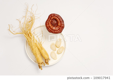 On a white background, fresh roots and slices of ginseng on white round ceramic dish decorated with lingzhi mushroom. Minimal concept for advertising product with rare herbal ingredient 109017941