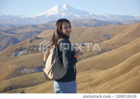A young brunette woman with a backpack stands against the backdrop of snow-capped Mount Elbrus and looks at the camera. 109018426