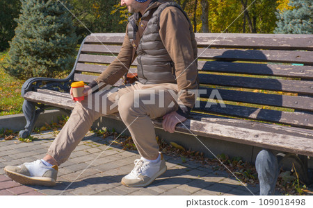 A young man sits on a bench in an autumn park, drinks coffee, rests, relaxes. Urban lifestyle concept. A young man sits on a bench in an autumn park, drinks coffee, rests, relaxes. Urban lifestyle concept. 109018498