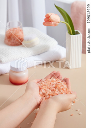 Female hand model with a lot of pink himalayan salt. A stack of white towels displayed with a flower pot and a beauty jar. Cosmetics, products for care and personal hygiene concept Female hand model with a lot of pink himalayan salt. A stack of white towels displayed with a flower pot and a beauty jar. Cosmetics, products for care and personal hygiene concept 109018699