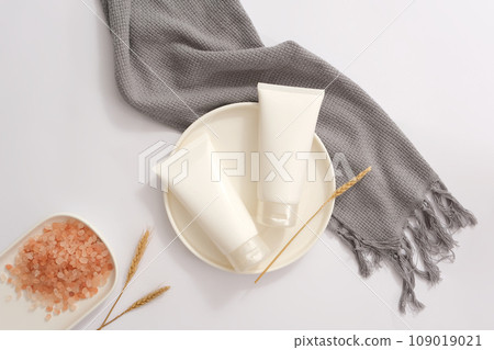 Round shaped dish featured two white tubes without label. A dish of pink himalayan salt displayed with a scarf. Daily self care routine concept  109019021