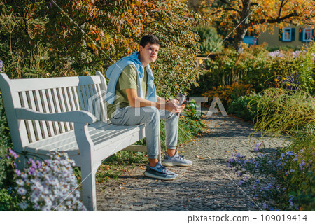 A Teenager Sits On A Bench In The Autumn Park Drinks Coffee From A Thermo Mug And Looks Into A Phone. Portrait Of Handsome Cheerful Guy Sitting On Bench Fresh Air Using Device Browsing Media Smm A Teenager Sits On A Bench In The Autumn Park Drinks Coffee From A Thermo Mug And Looks Into A Phone. Portrait Of Handsome Cheerful Guy Sitting On Bench Fresh Air Using Device Browsing Media Smm 109019414