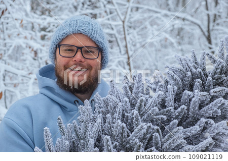 Having in his hands a snowy Christmas tree, a cheerful man in blue smiles 109021119