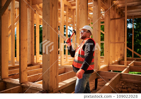 Carpenter constructing wooden frame house. Bearded man with glasses is hammering nails into the structure, wearing protective helmet and construction vest. Concept of modern ecological construction. Carpenter constructing wooden frame house. Bearded man with glasses is hammering nails into the structure, wearing protective helmet and construction vest. Concept of modern ecological construction. 109022028