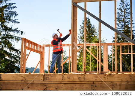 Wooden frame-built home is being erected by carpenter near forest. Bearded man wearing glasses driving in nails with hammer while sporting a safety helmet and a construction vest. Wooden frame-built home is being erected by carpenter near forest. Bearded man wearing glasses driving in nails with hammer while sporting a safety helmet and a construction vest. 109022032
