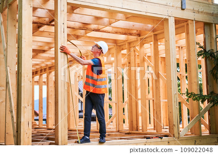 Carpenter building wooden-framed house. Man measures distances with measuring tape, donned in work attire and protective helmet. Principles of modern environmentally conscious construction. 109022048