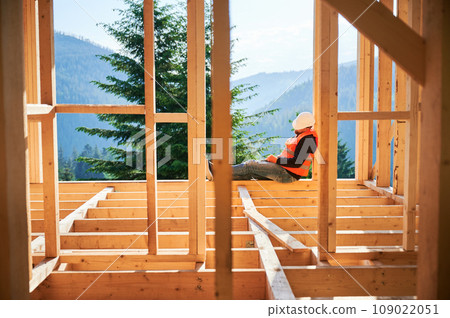 Carpenter constructing two-story wooden frame house near the forest. Man taking break from work, dressed in protective helmet and orange vest. Concept of lunchtime nap at construction site. 109022051
